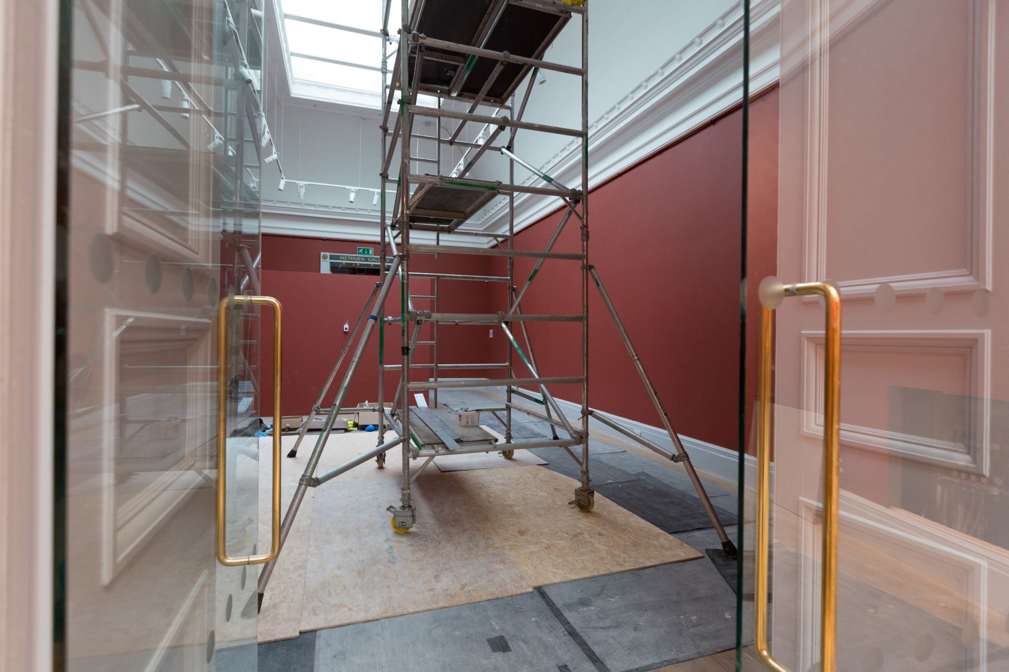 Glass double doors lead into a gallery space with red walls and scaffolding in the centre 
