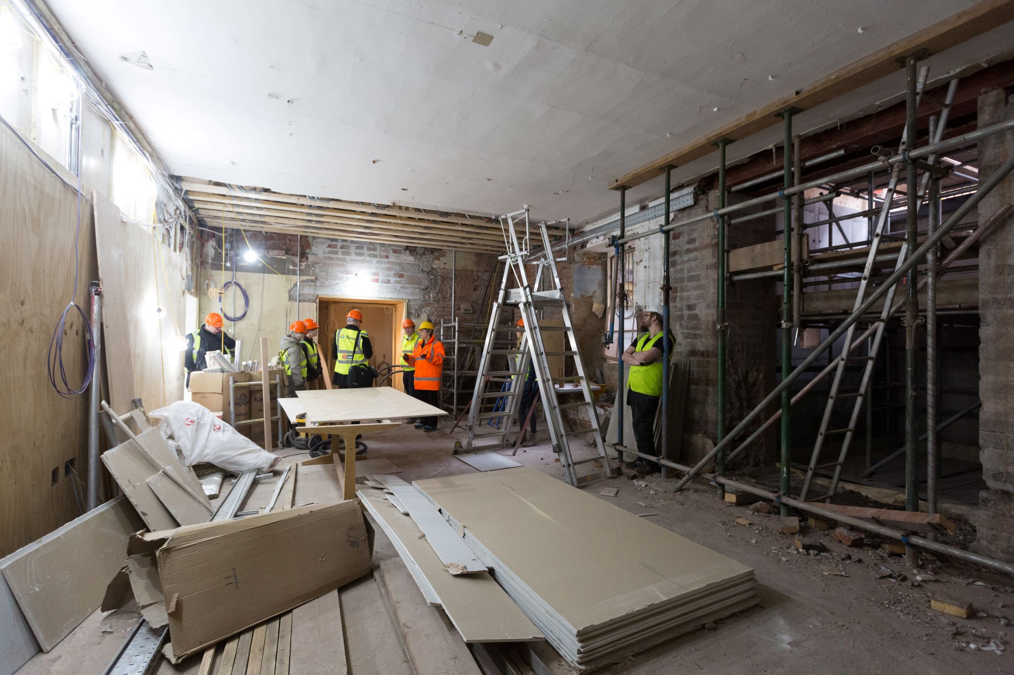 A room with scaffolding and building materials & people in high vis vests & safety helmets at the far side of the space 