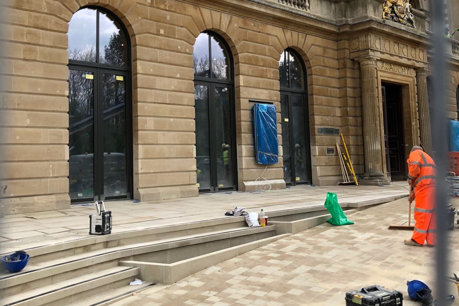 The entrance to the RWA, with large glass windows, paving in the foreground and someone in an orange high vis jacket sweeping at the side 