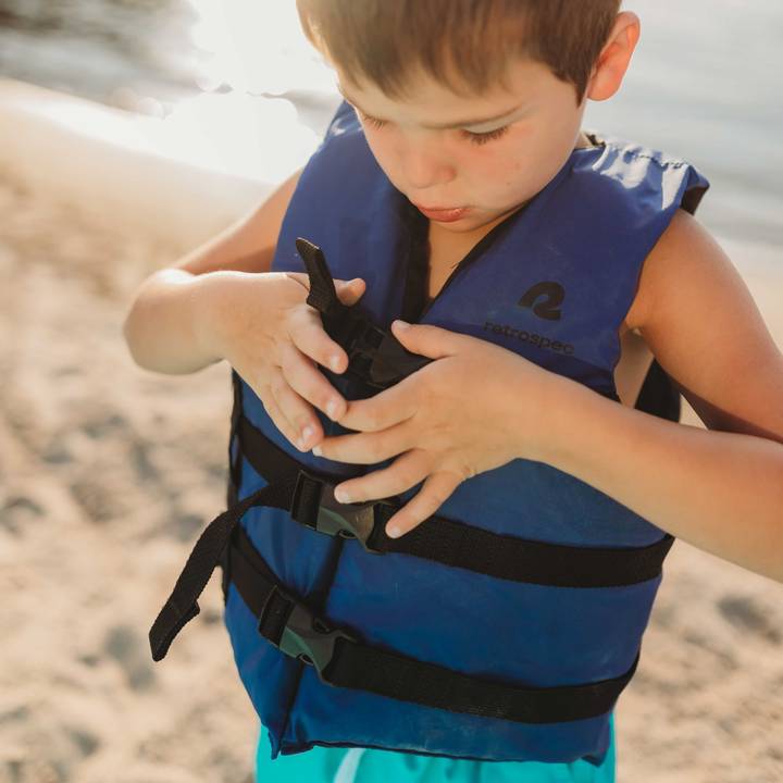 A young boy fastening the buckle straps on his retrospec Bobbin’ Youth Life Jacket in Harborside Blue while standing on a sandy beach at golden hour.