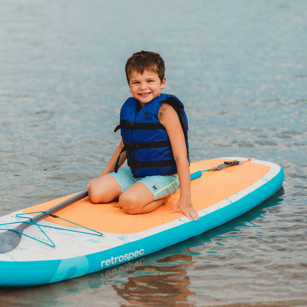 A smiling boy wearing a  retrospec Bobbin’ Youth Life Jacket in Harborside Blue sits cross-legged on a retrospec Weekender Nano Kids Inflatable Stand Up Paddle Board 8’6" floating on calm water, with a paddle resting across the board.