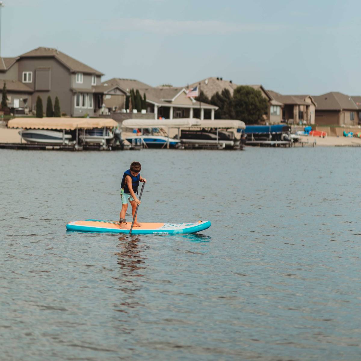 A young boy wearing a retrospec Bobbin’ Youth Life Jacket in Harborside Blue paddles standing on a retrospec Weekender Nano Kids Inflatable Stand Up Paddle Board 8’6" on a calm lake, with lakeside homes, boat docks, and a hazy sky visible in the background.