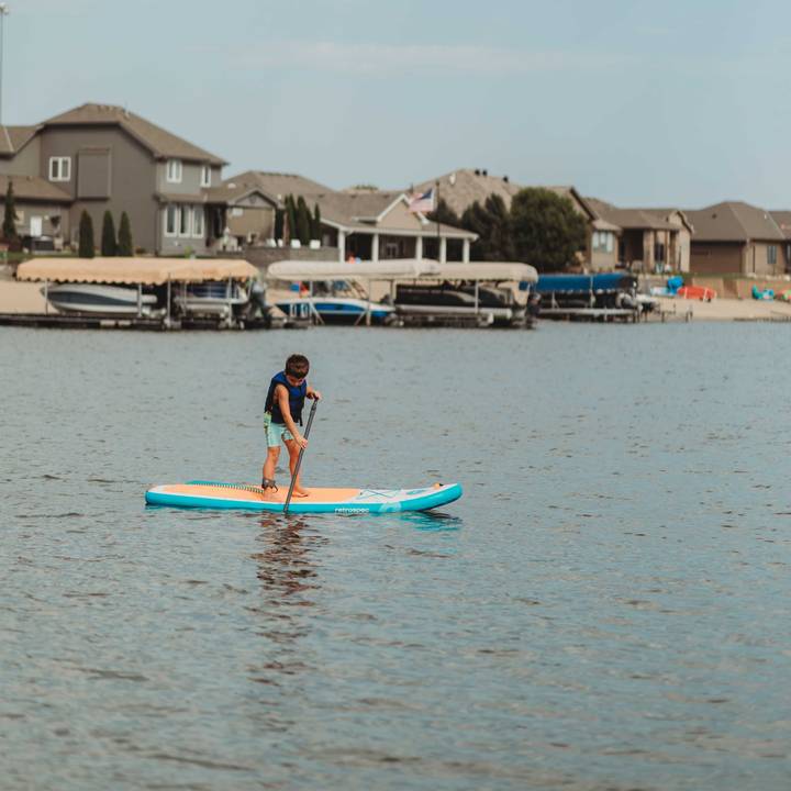 A young boy wearing a retrospec Bobbin’ Youth Life Jacket in Harborside Blue paddles standing on a retrospec Weekender Nano Kids Inflatable Stand Up Paddle Board 8’6" on a calm lake, with lakeside homes, boat docks, and a hazy sky visible in the background.