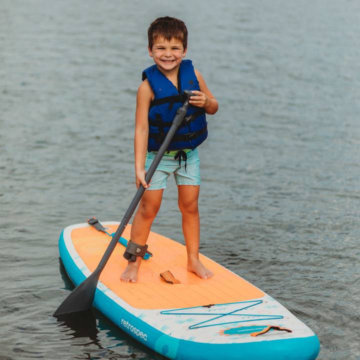 A smiling child standing on a retrospec inflatable Paddle Board on calm water, wearing a retrospec Bobbin’ Youth Life Jacket in Harborside Blue and holding a paddle.

