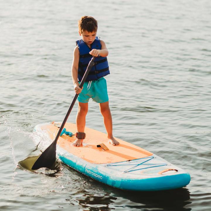 A smiling child standing on a retrospec inflatable Paddle Board on calm water, wearing a retrospec Bobbin’ Youth Life Jacket in Harborside Blue and holding a paddle.
