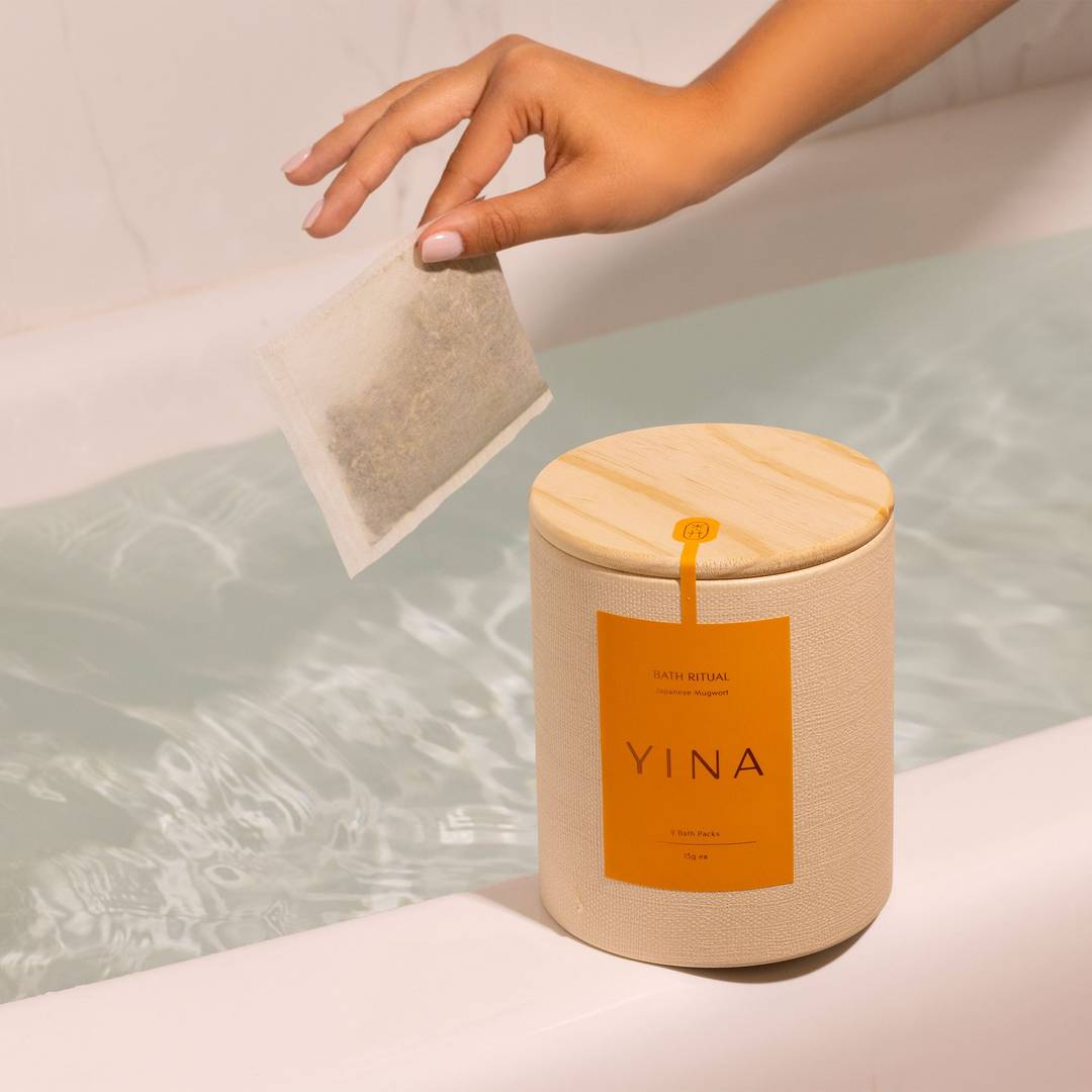 A hand holding a mugwort bath pack over a bath tub filled with warm water with a canister of YINA Bath Ritual canister product on the ledge of the tub.