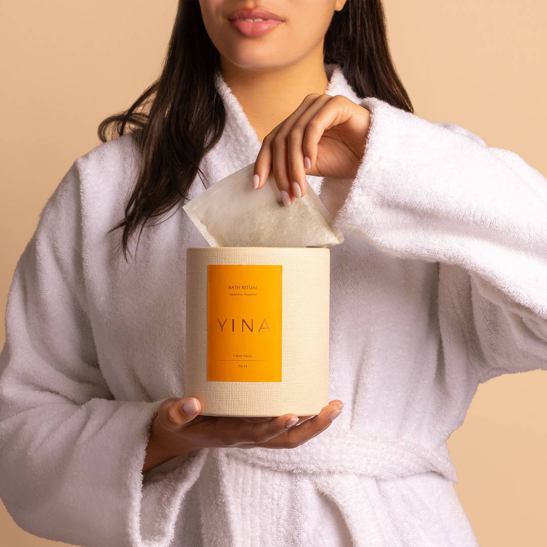 Woman in bath robe holding a canister with one hand and puling out a mugwort bath pack with the other.