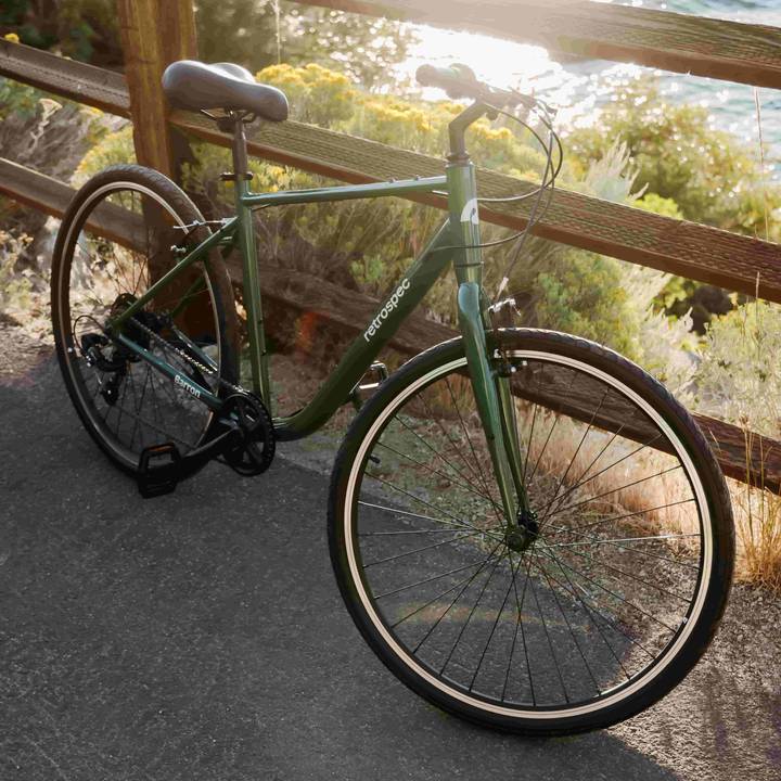 A green Barron Comfort Hybrid 7 Speed Bike parked on a paved path by a wooden fence, with scenic greenery and a body of water in the background.