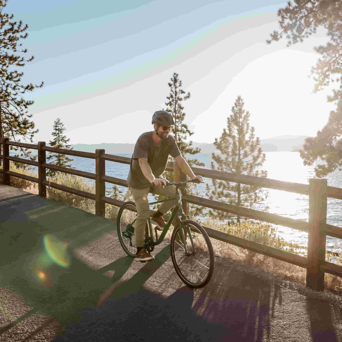 Person cycling on a path by a lake with sunlight reflecting off the water and pine trees in the background.