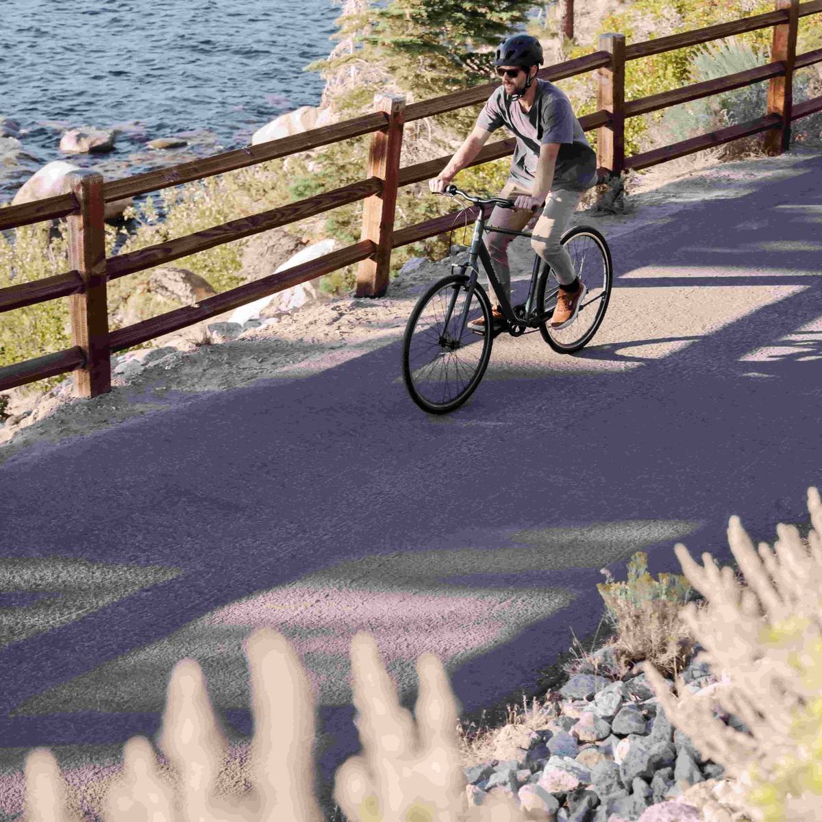 Person cycling on a paved path beside a body of water, with a wooden fence and surrounding vegetation.
