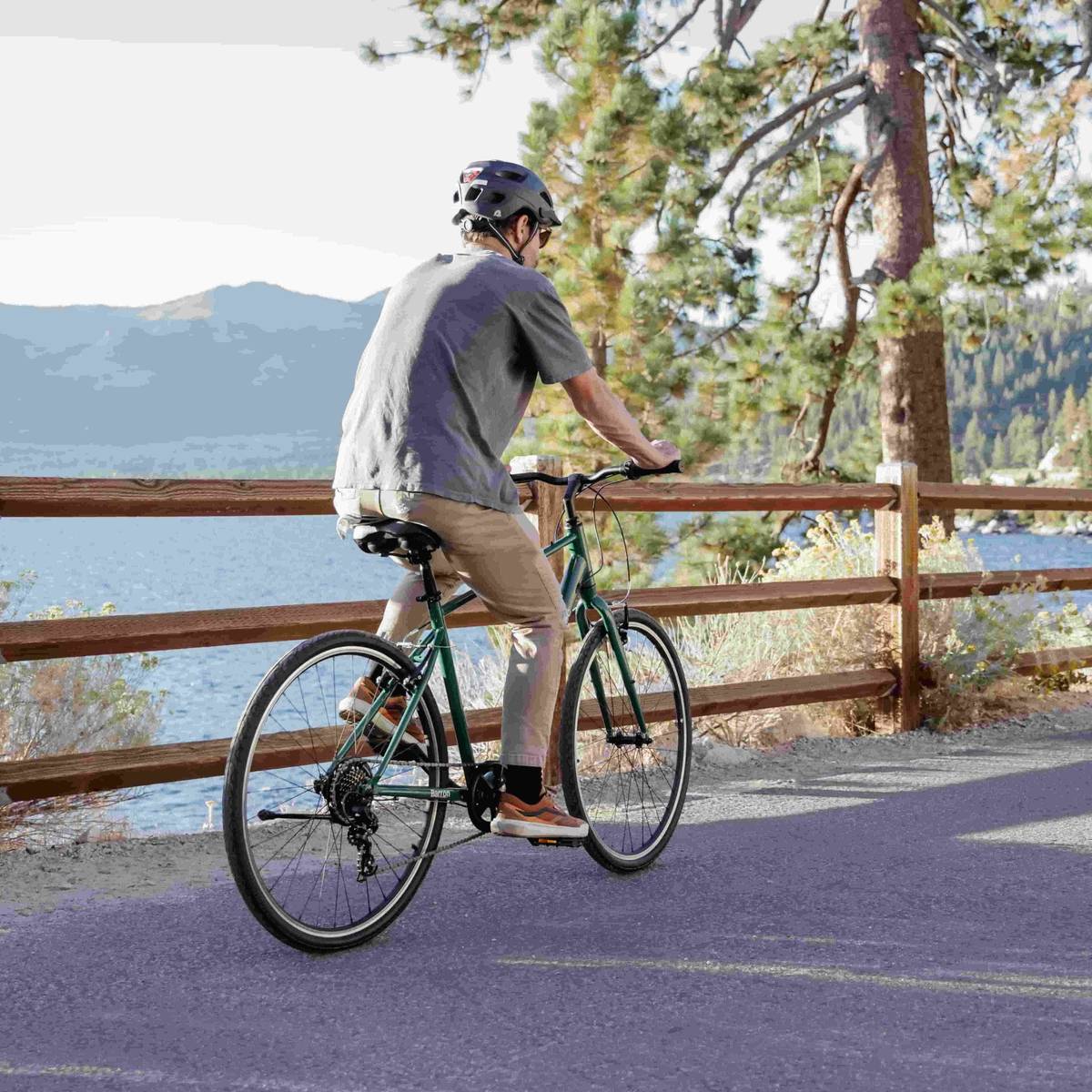 A person cycling on a path near a lake, with mountains and pine trees in the background.