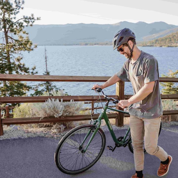 Person walking a Barron Comfort Hybrid 7 Speed Bike near a lake with mountains in the background.