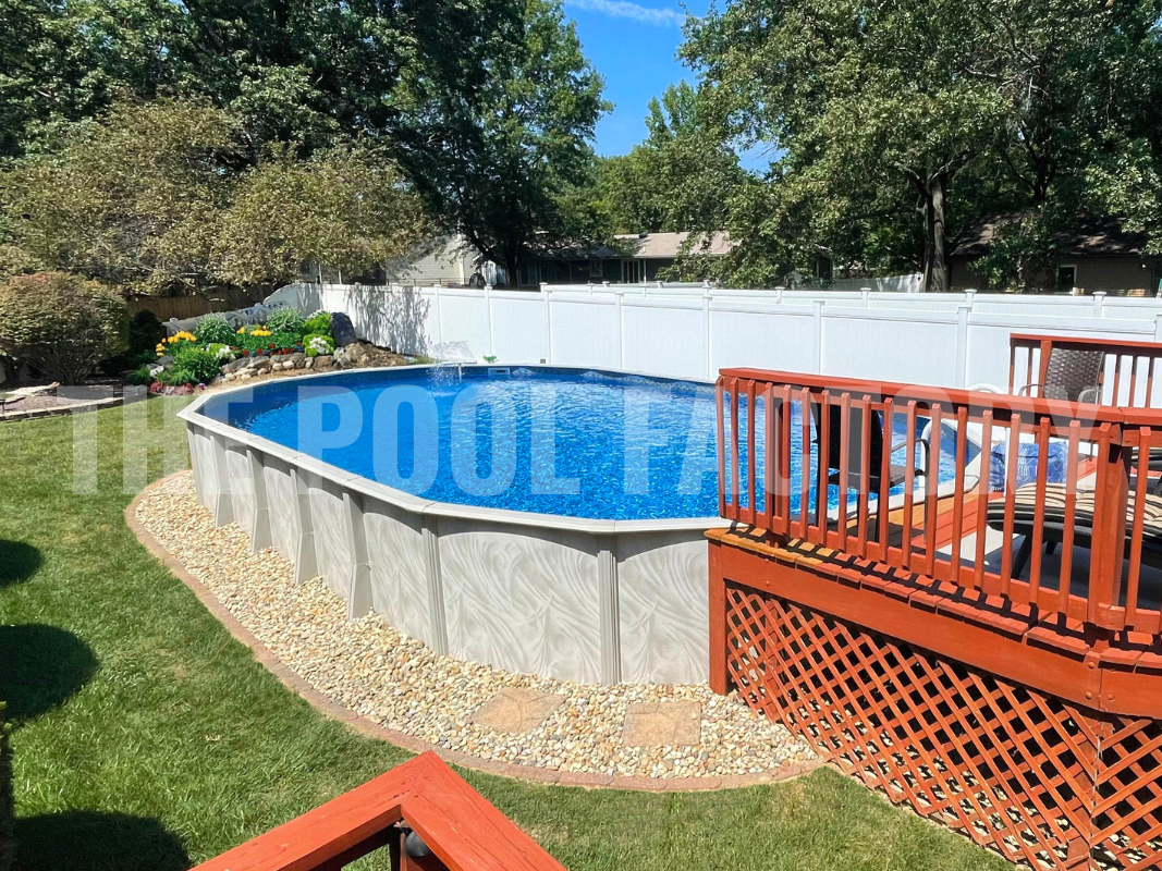 Oval above ground swimming pool with partial deck, surrounded by trees, plants and flower beds