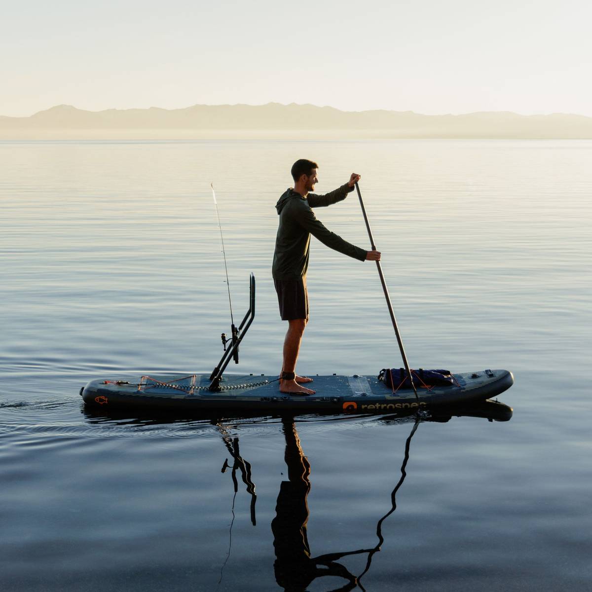 A person stands while paddling their June Inflatable Fishing Paddle Board Kayak Hybrid with the fishing rod holder attached to the board in calm water while the sun it setting.