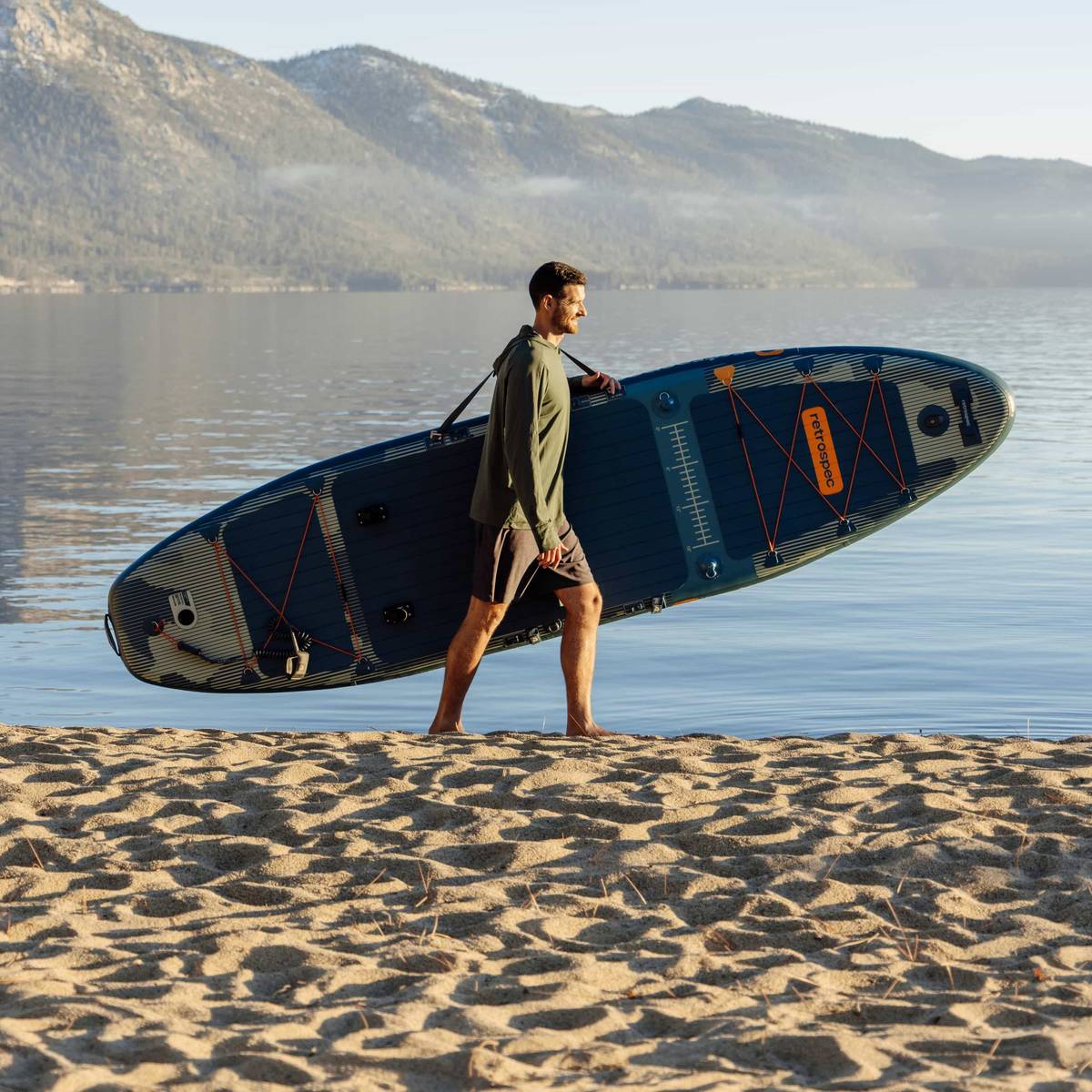 A person using a carry strap to walk with their inflatable fishing paddle board kayak hybrid on the sand at the beach with the water and mountains in the background.
