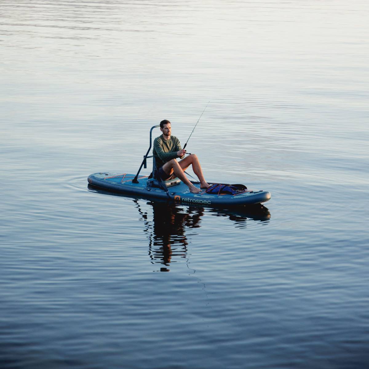 A person sitting on the June Inflatable Fishing Paddle Board Kayak Hybrid, fishing in calm water with reflections, surrounded by a serene outdoor setting.