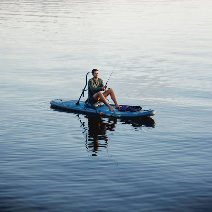 A person sitting on the June Inflatable Fishing Paddle Board Kayak Hybrid, fishing in calm water with reflections, surrounded by a serene outdoor setting.