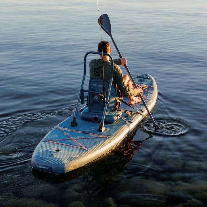 A person sitting on the June Inflatable Fishing Paddle Board Kayak Hybrid, paddling in calm water with reflections, surrounded by a serene outdoor setting.