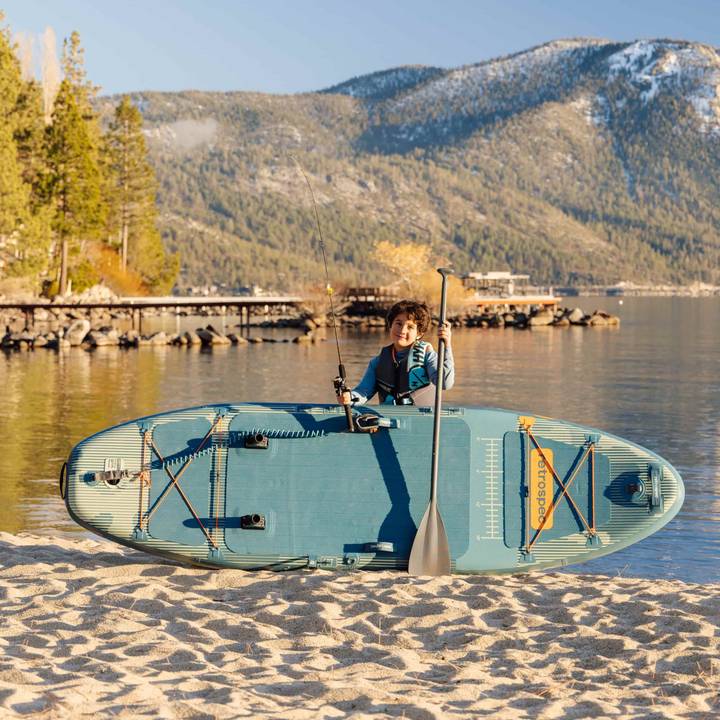 A child rests with their June Nano Kids Inflatable Fishing Paddle Board Kayak Hybrid and paddle on the sand with a calm body of water in the background.