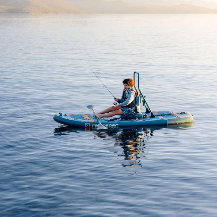 A child fishes while sitting on a June Nano Kids Inflatable Stand Up Paddle Board Kayak Hybrid with 'retrospec' branding on a calm body of water.