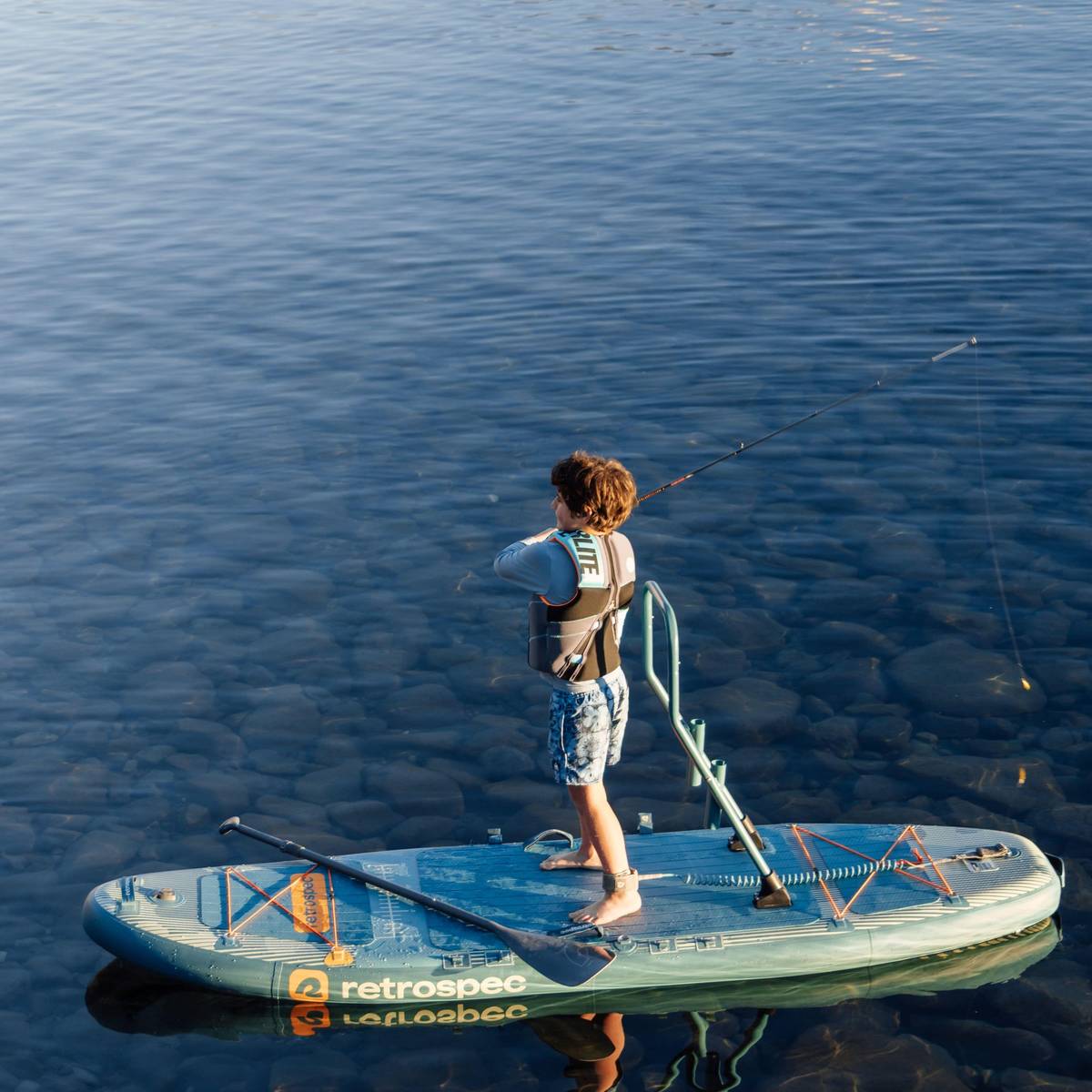 A child casts while standing on a June Nano Kids Inflatable Fishing Stand Up Paddle Board Kayak Hybrid with 'retrospec' branding on a calm body of water.