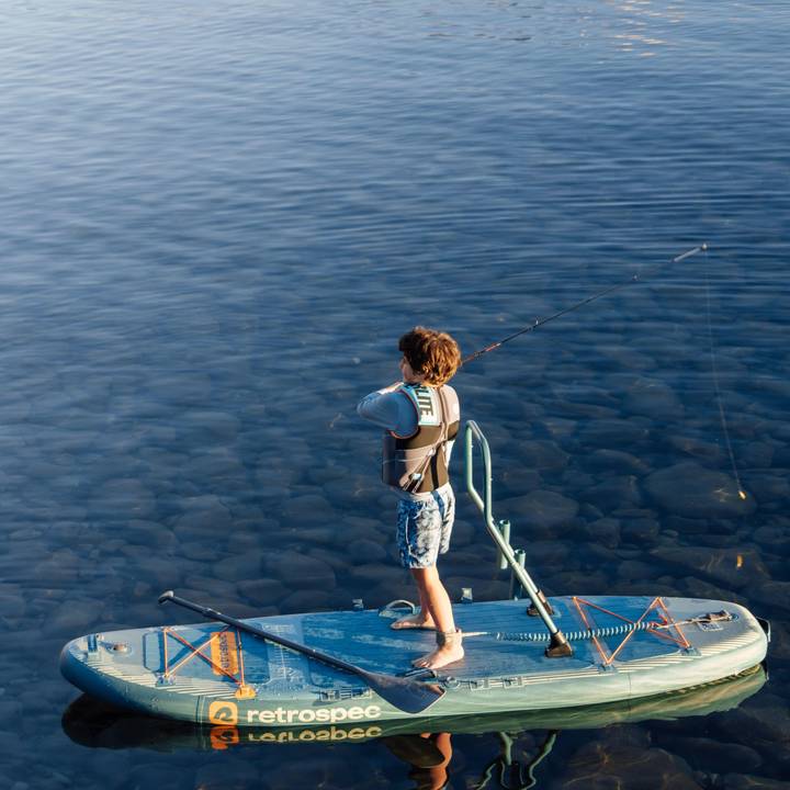 A child casts while standing on a June Nano Kids Inflatable Fishing Stand Up Paddle Board Kayak Hybrid with 'retrospec' branding on a calm body of water.