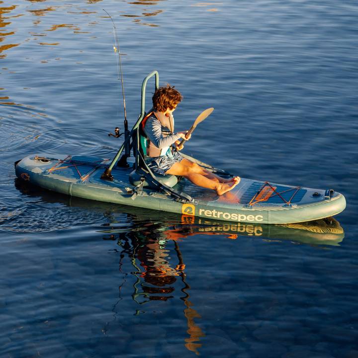 A child paddles while sitting on a June Nano Kids Inflatable Stand Up Paddle Board Kayak Hybrid with 'retrospec' branding on a calm body of water.