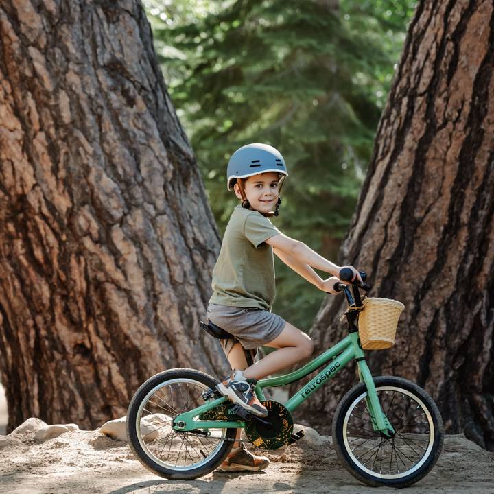 A child on a green Koda 16" Kids Bike with a basket, wearing a helmet, riding on a dirt path between two large trees in a forest setting.