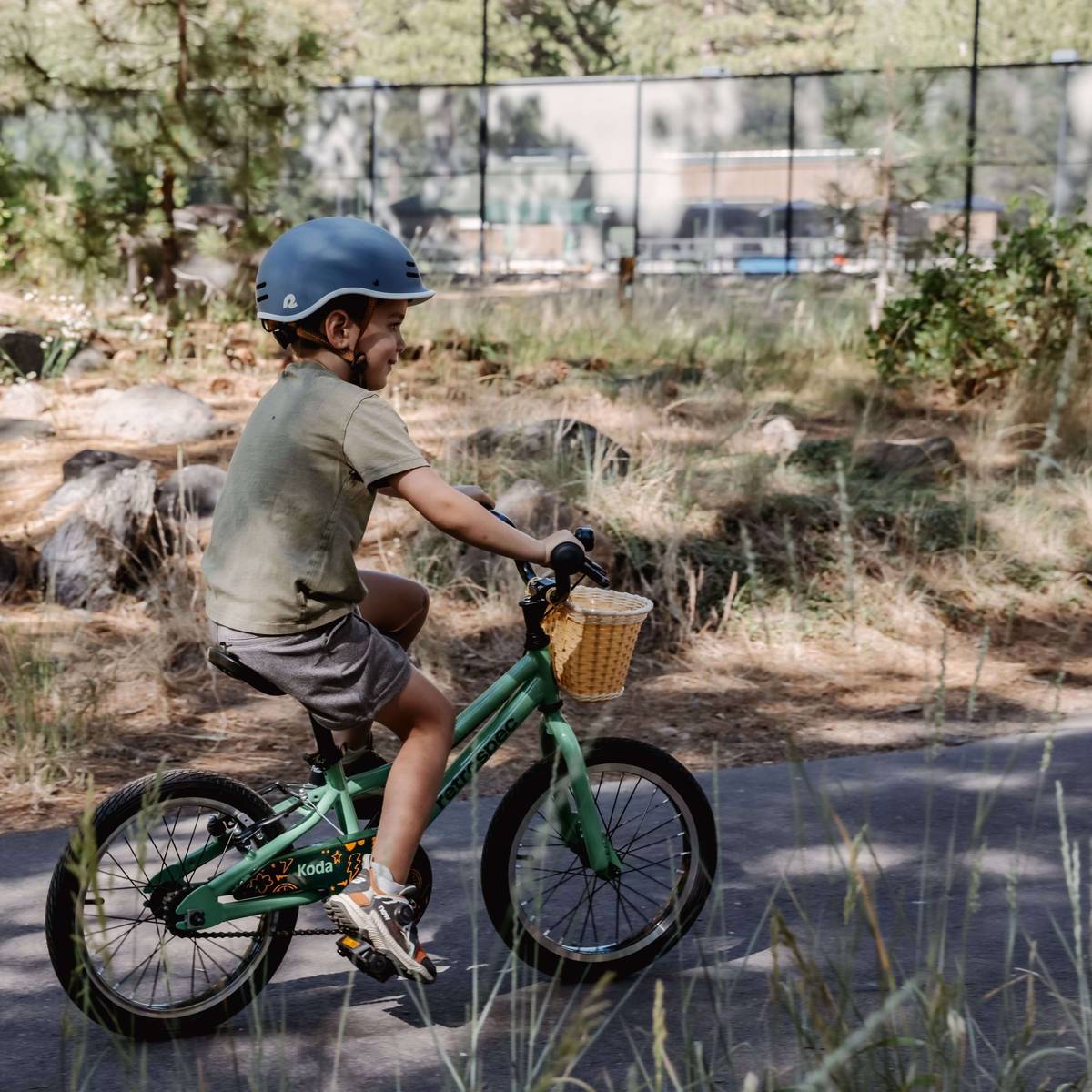 A child rides a green Koda 16" Kids Bike with a wicker basket, wearing a blue helmet, surrounded by trees and grassy terrain on a sunny day.