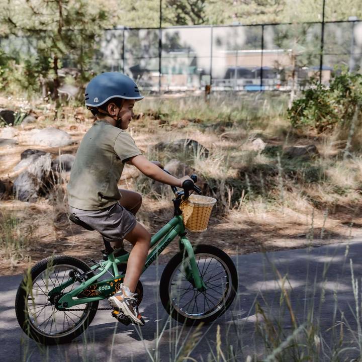 A child rides a green Koda 16" Kids Bike with a wicker basket, wearing a blue helmet, surrounded by trees and grassy terrain on a sunny day.