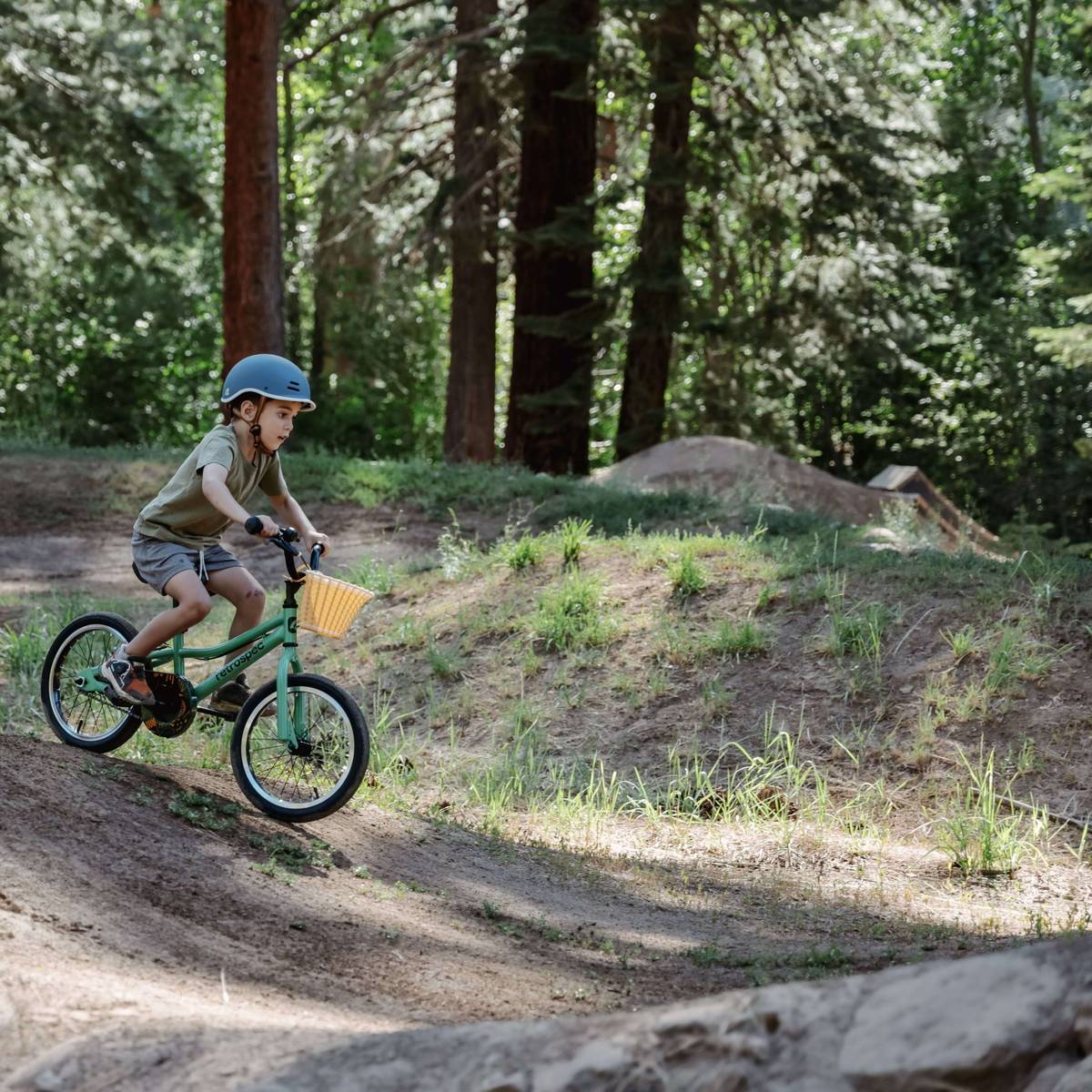 A child rides a green Koda 16" Kids Bike down a slope in a wooded area, wearing a helmet and shorts, surrounded by lush greenery and dirt paths.