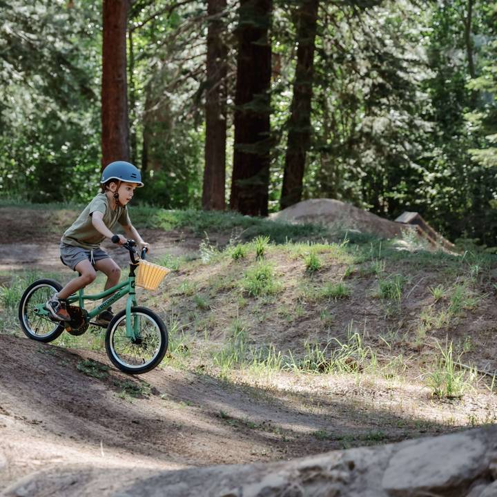 A child rides a green Koda 16" Kids' Bike down a slope in a wooded area, wearing a helmet and shorts, surrounded by lush greenery and dirt paths.