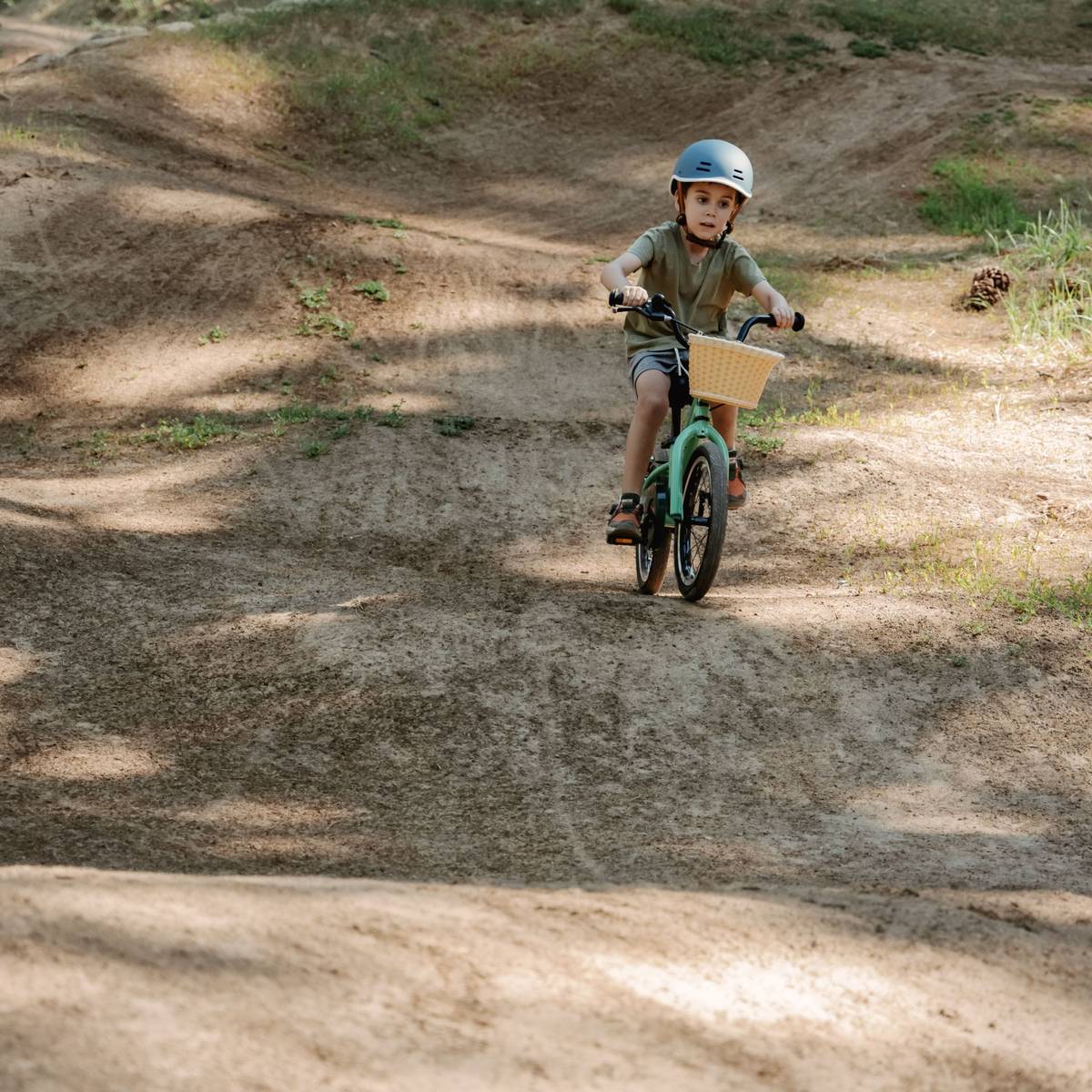 A child is riding a green Koda 16" Kids Bike with a wicker basket on a dirt path, surrounded by grass and uneven terrain.