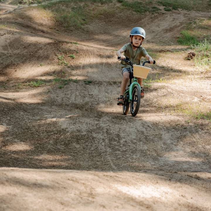 A child is riding a green Koda 16" Kids Bike with a wicker basket on a dirt path, surrounded by grass and uneven terrain.