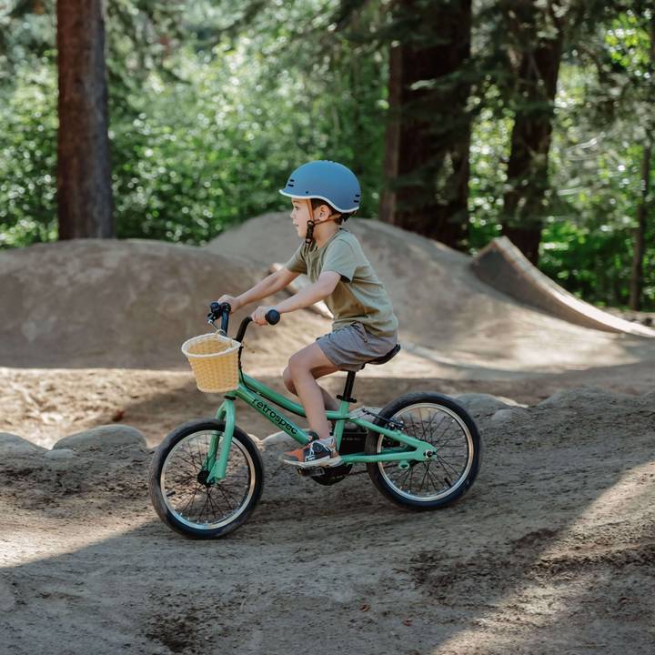 A child rides a green Koda 16" Kids Bike with a basket, navigating a dirt track surrounded by tall trees in a sunny forest.