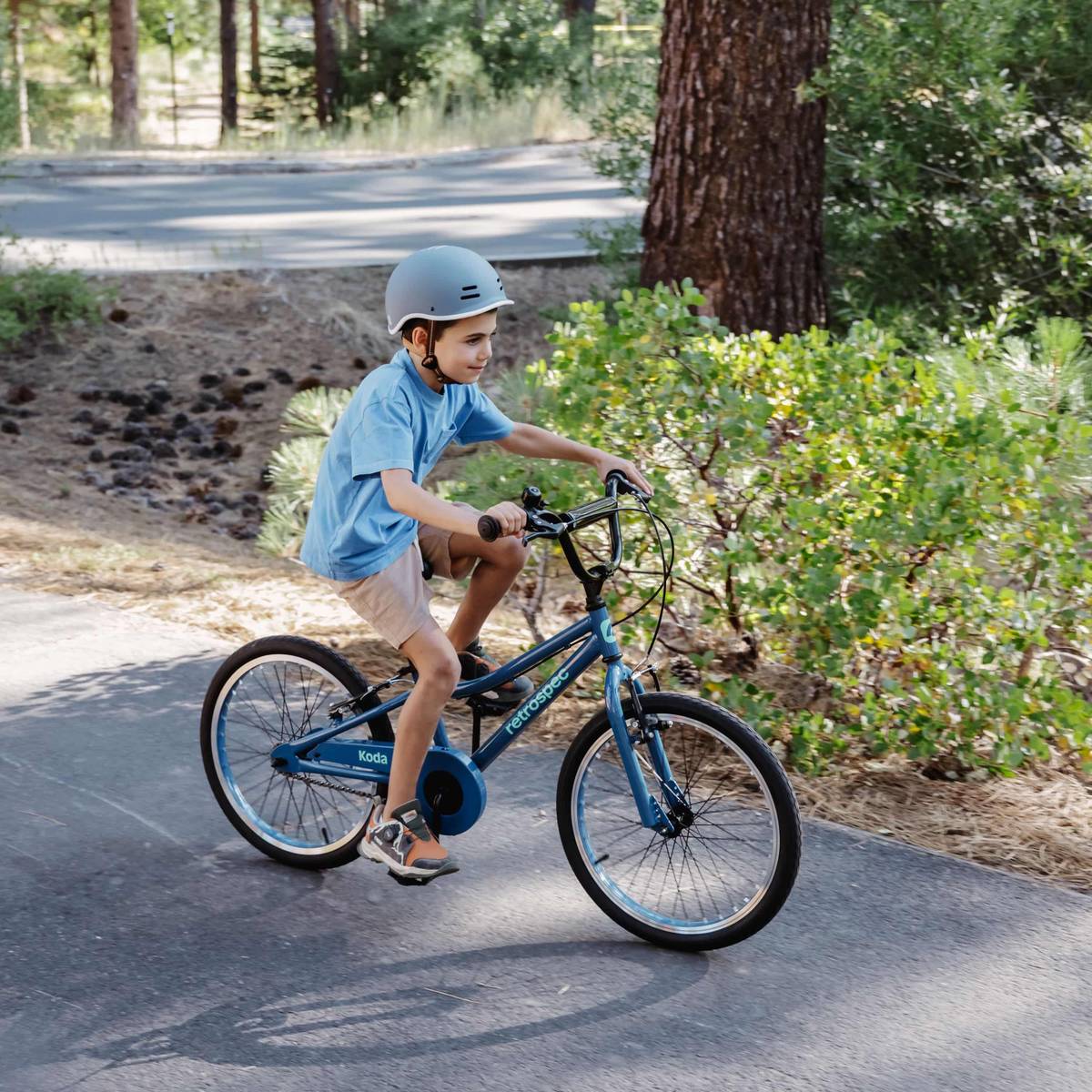 A young boy joyfully riding a blue Koda 20" Kids Bike on a sunny day, wearing a helmet and a blue t-shirt.