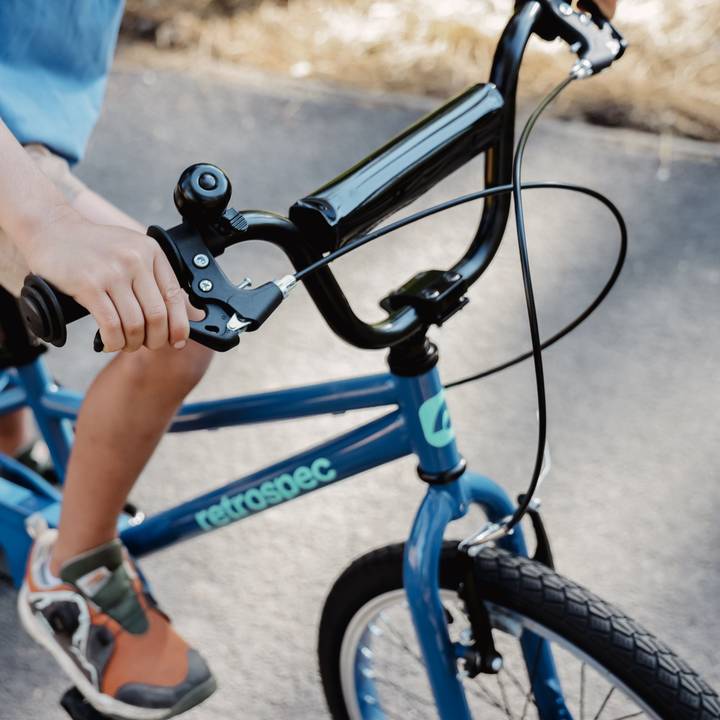 Detailed view of a child's blue Koda 20" Kids Bike handlebars and hand brake while riding on a paved path.