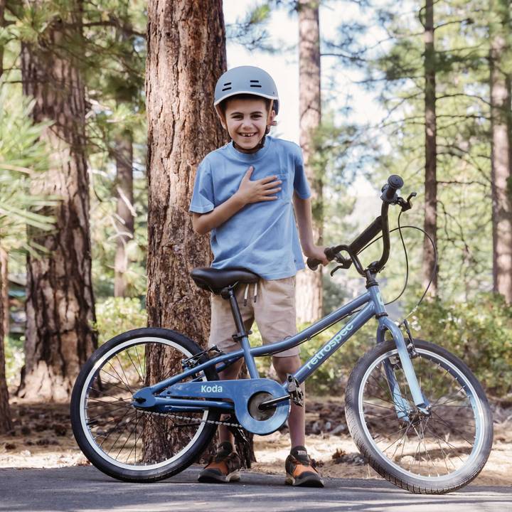 A young boy in a helmet stands next to a blue Koda 20" Kids Bike, smiling and prepared for cycling.