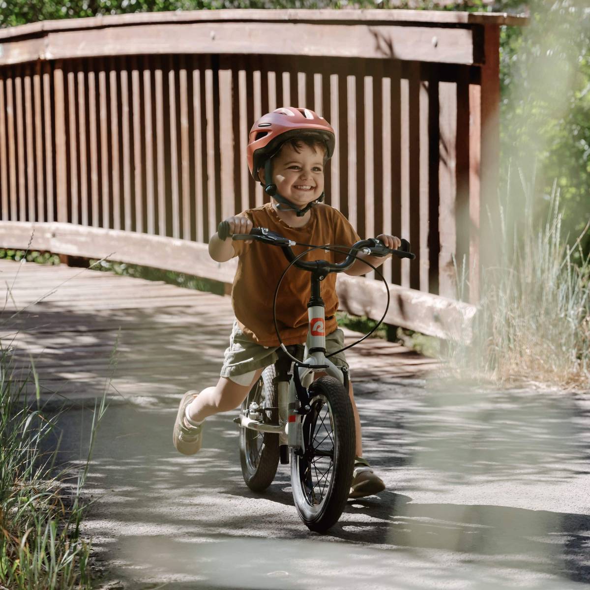 A young child rides a Cub Plus XL 14" Kids' Balance Bike across a bridge, enjoying the outdoors on a sunny day.