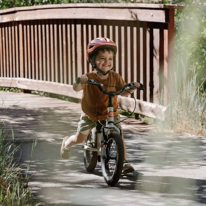 A young child rides a Cub Plus XL 14" Kids' Balance Bike across a bridge, enjoying the outdoors on a sunny day.