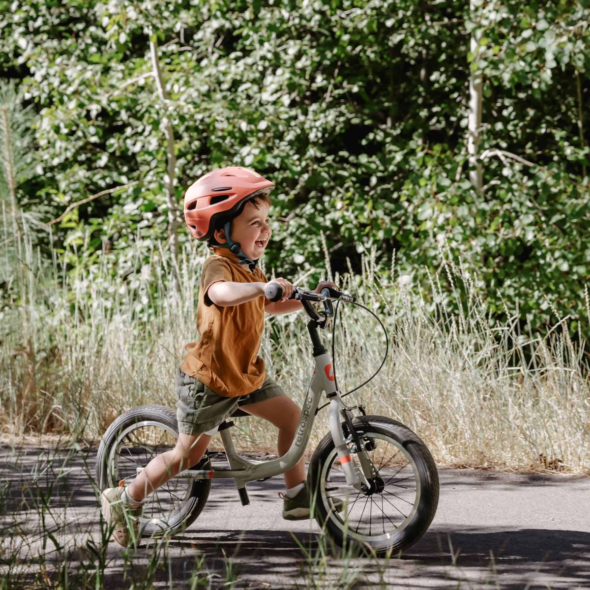 A young child in a helmet rides a Cub Plus XL 14" Kids' Balance Bike along a scenic trail surrounded by trees and greenery.