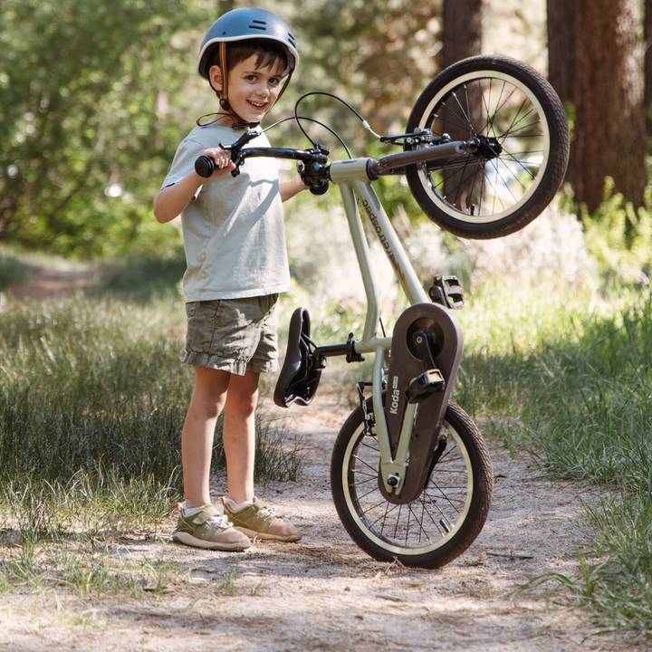 A child in casual attire balances a Koda Plus 16" Kids bike on its back wheel, surrounded by trees and grassy terrain on a sunny day.