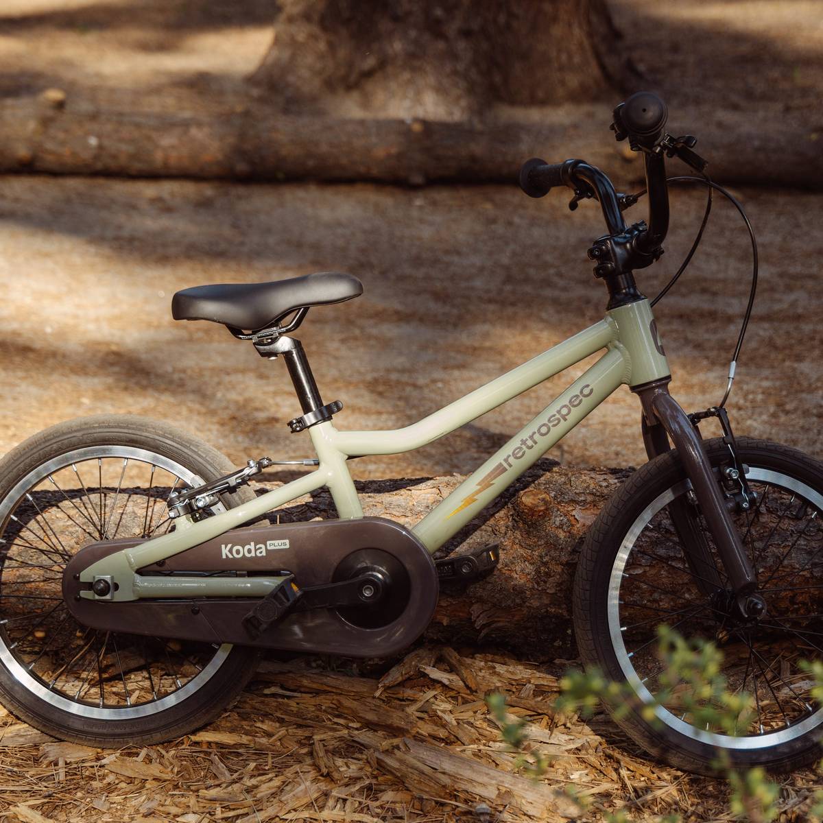 A vintage-style green Cub Plus 16" Kids Bike rests on a log, surrounded by natural wood chips and foliage, showcasing its sleek design.
