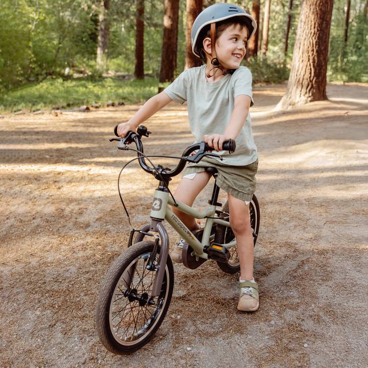 A Koda Plus 16" bike in soft green, resting against a log in a woodland setting, showcasing its sleek design and sturdy frame.