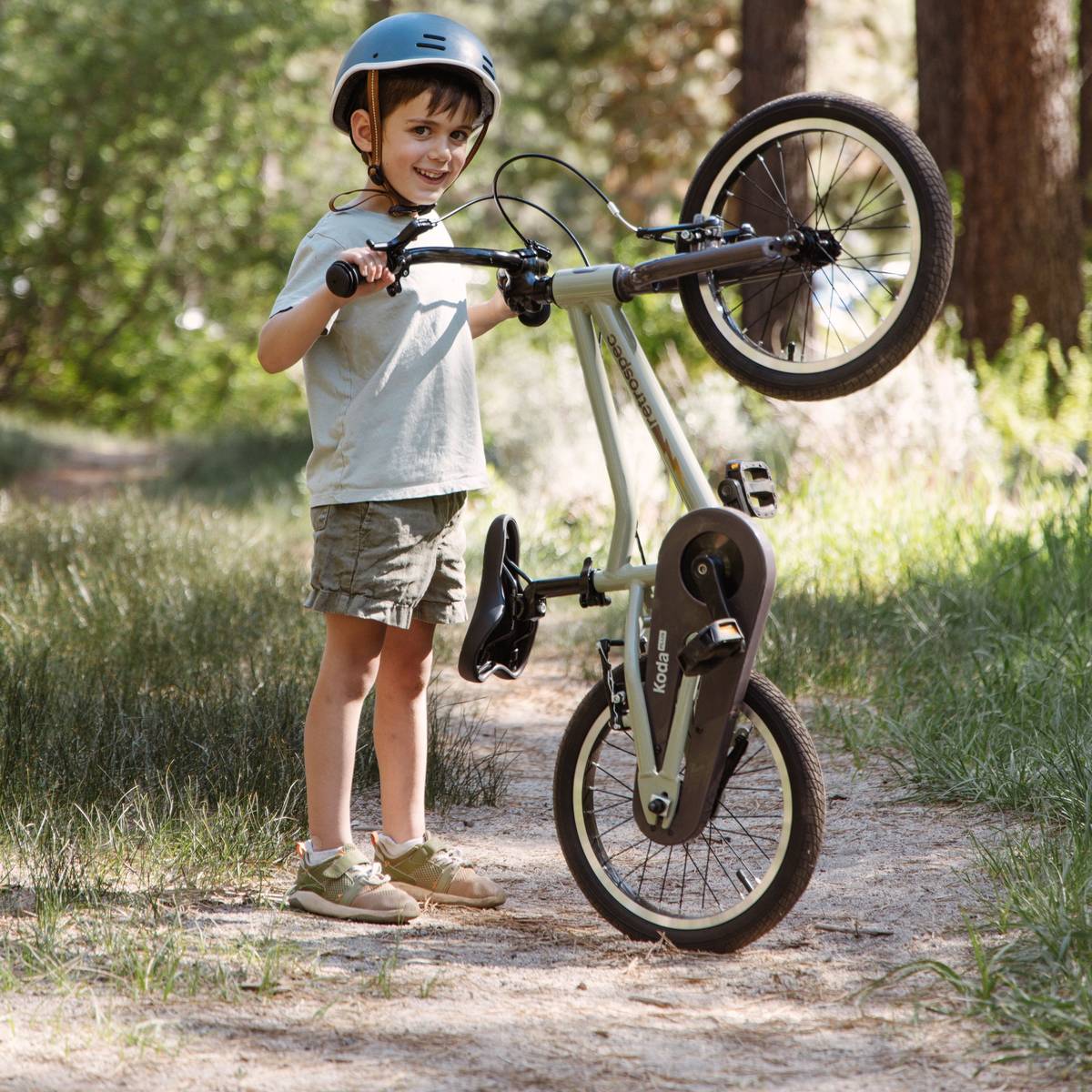 A child in casual attire balances a Koda Plus 16" Kids bike on its back wheel, surrounded by trees and grassy terrain on a sunny day.