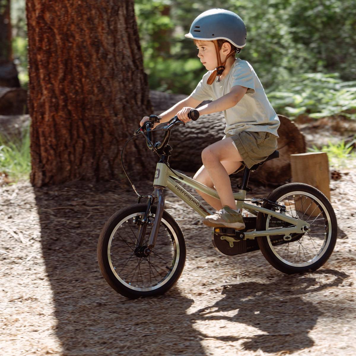 A child rides a Koda Plus 16" Kids Bike on a forest path, wearing a helmet and casual clothing, surrounded by trees and natural scenery.