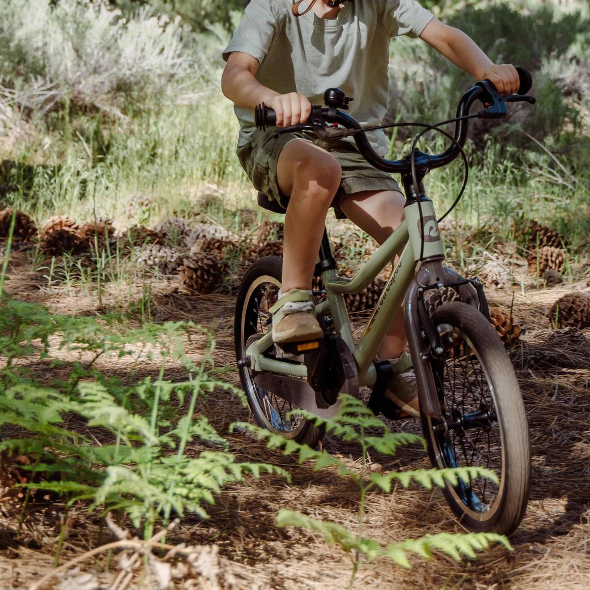 A child rides an olive-green Koda Plus 16" Kids Bike through a forested area with ferns and pine cones scattered on the ground.