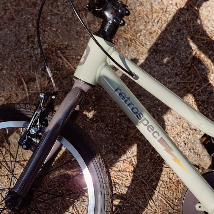 Close-up of a Koda Plus 16" Kids Bike frame, showcasing its pale green color, black handlebars, and sturdy wheel against a gravel background.