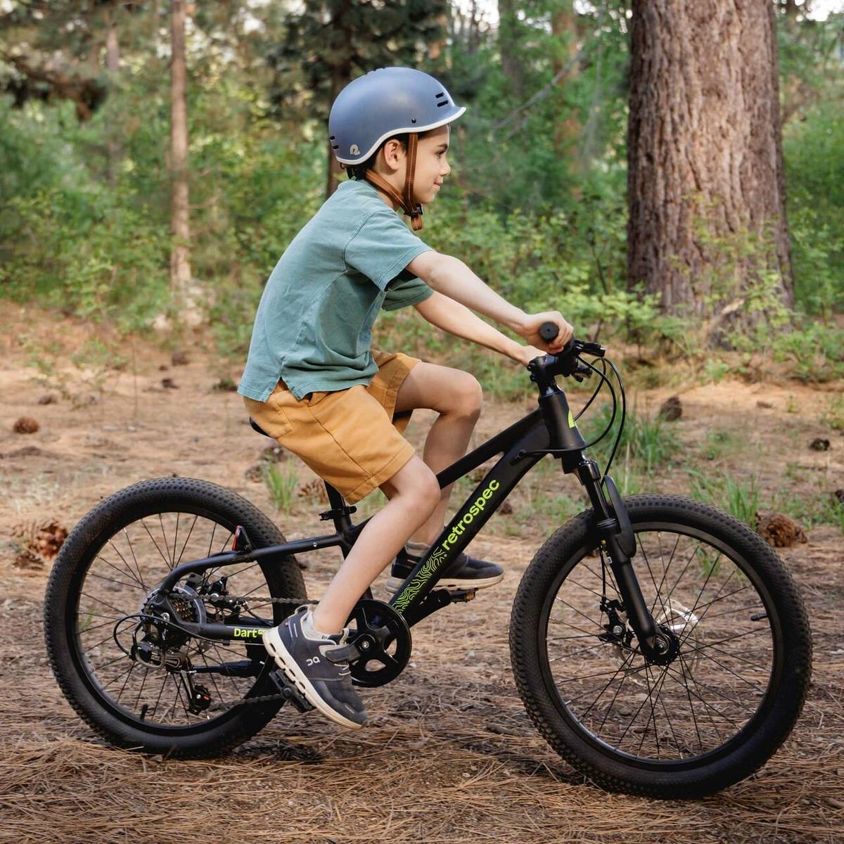 A child rides a black Dart Plus 20in  bicycle in a forest, wearing a blue Remi helmet, a green shirt, and shorts, surrounded by trees and pine needles.