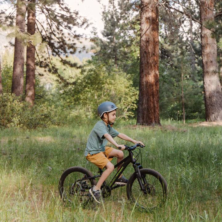 A child wearing a Remi Kids Bike Helmet rides a black Dart Plus 20" Kids Bike 7 Speed through a grassy area surrounded by tall trees.